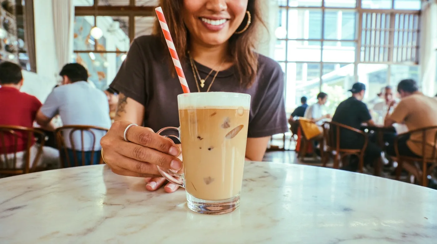 Woman with a tall glass of Thai iced coffee at a sunlit Bangkok food hall table