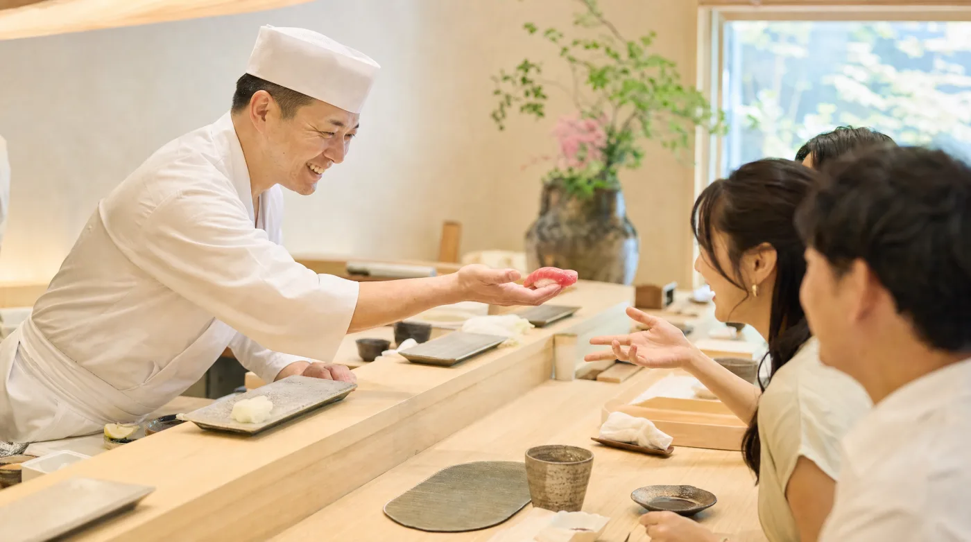 Sushi chef handing a piece of nigiri across a bright hinoki wood counter to seated guests